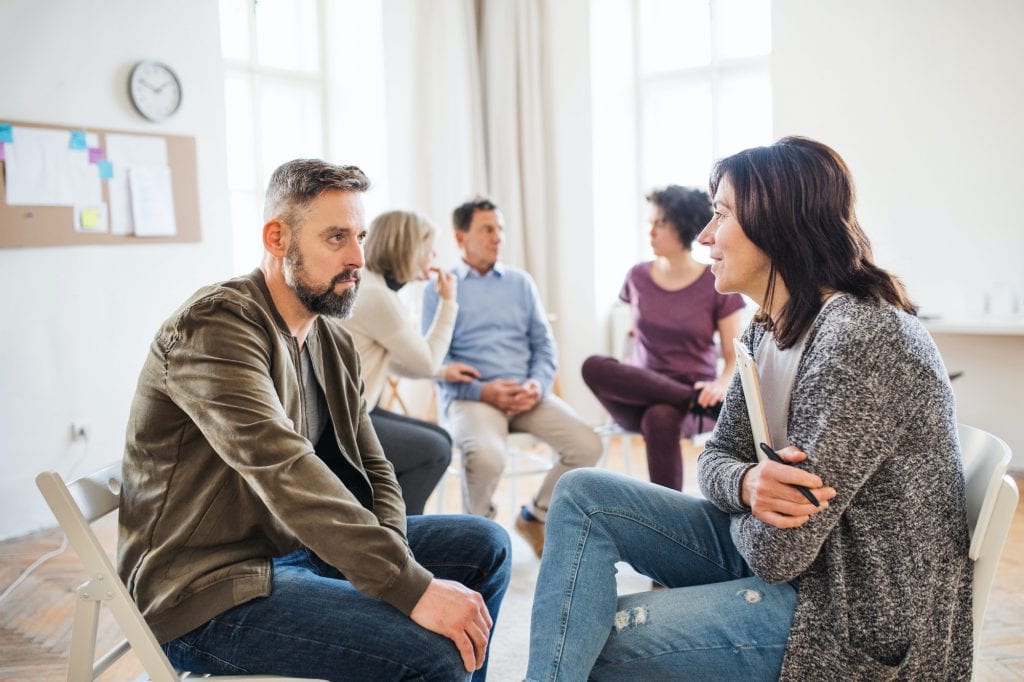 Senior counselor with clipboard talking to a man during group therapy.
